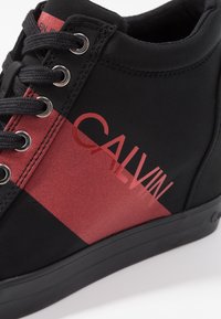 Černé látkové vysoké tenisky s červeným diagonálním pruhem a textem "CALVIN", kovovými očky a texturovanými detaily na svršku.