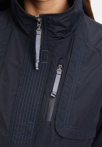 Laivastonsininen takki korkealla kauluksella, jossa on viistosti ommeltuja saumoja, kaksinkertaiset vetoketjulliset taskut ja raidalliset vetoketjunvedot edessä.