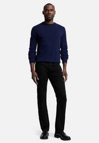 Marine blauwe wollen crewneck trui, zwarte jeans en zwarte veterschoenen gedragen door een persoon die op een effen achtergrond staat.