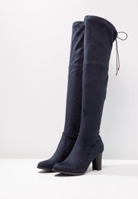 Botas altas hasta el muslo de ante azul marino con un diseño ajustado, punta puntiaguda y tacón grueso. Cuentan con un lazo ajustable en la parte superior.