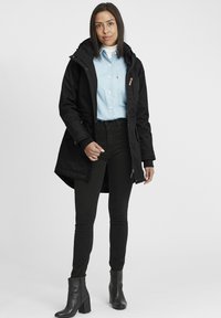 Parka noire avec capuche, deux poches avant et fermeture éclair. Porté sur une chemise bleu clair et un jean skinny noir avec des bottes montantes.