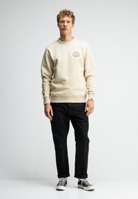 Beige sweatshirt met geribbelde manchetten en zoom, met een kleine grafische afbeelding op de borst. Gepaard met zwarte jeans en zwart-witte sneakers.