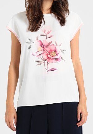 Femme portant un chemisier blanc à manches courtes avec un imprimé floral rose et gris, associé à un pantalon bleu marine foncé.