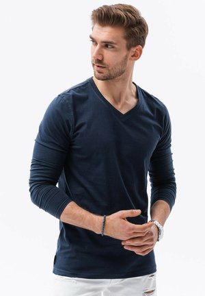 Marineblå langærmet V-hals t-shirt lavet af blødt bomuld, med et tætsiddende design og synlige sømdetaljer. Bæres med hvide slidte jeans.