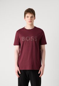Burgundská bavlnená tričko s výrazným logom "BOSS" v štruktúrovanom šedivom vzore. Krátke rukávy, okrúhly výstrih a voľný strih.