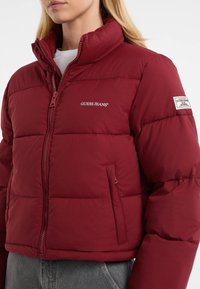 Primo piano di una persona che indossa una giacca a vento rossa di Guess Jeans con tasche con zip e loghi brandizzati sul petto e sulle maniche.