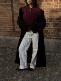 Palton negru din blană artificială peste un tricou polo burgund, asortat cu blugi albi cu croială largă și saboți maro pe o suprafață de piatră cubică.