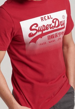 Czerwona koszulka z białym prostokątnym logo z napisem "REAL SuperDry" i japońskimi znakami, noszona przez osobę z jedną ręką w kieszeni.