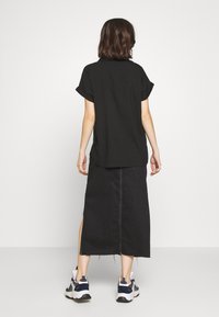 Chemise noire à manches courtes associée à une longue jupe en denim noir avec des fentes latérales et des coutures blanches. Portée avec des baskets noires et blanches.