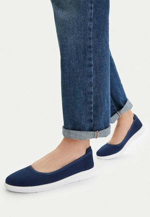 Osoba nosząca podwinięte niebieskie dżinsy i granatowe wsuwane buty z białymi wyściełanymi podeszwami, stojąca na jasnej powierzchni.