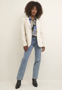 Blazer crème avec un bouton, blouse à motifs bleu et beige, jeans bleu clair, bottines noires à talons et cheveux lâches et bouclés.