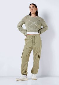 Γυναίκα φορώντας ένα ανοιχτό πράσινο πλεκτό πουλόβερ και ταιριαστά cargo παντελόνια συνδυασμένα με λευκά αθλητικά παπούτσια, στέκεται με τα χέρια στη μέση μπροστά από ένα απλό φόντο.