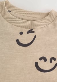 Beige bomuldsweater med ribbestrikket krave, prydet med to sorte smiley-ansigt-prints. Blød tekstur med et afslappet design.