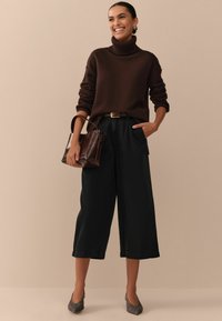 Dunkelbrauner Rollkragenpullover, hochgeschnittene schwarze weit geschnittene Hose, braune Handtasche und strukturierte graue Absätze. Neutrale Hintergrund.