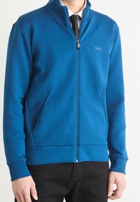 Blå zip-up jacka i mjukt tyg, med hög krage, två sidofickor och en diskret logotyp på bröstet.