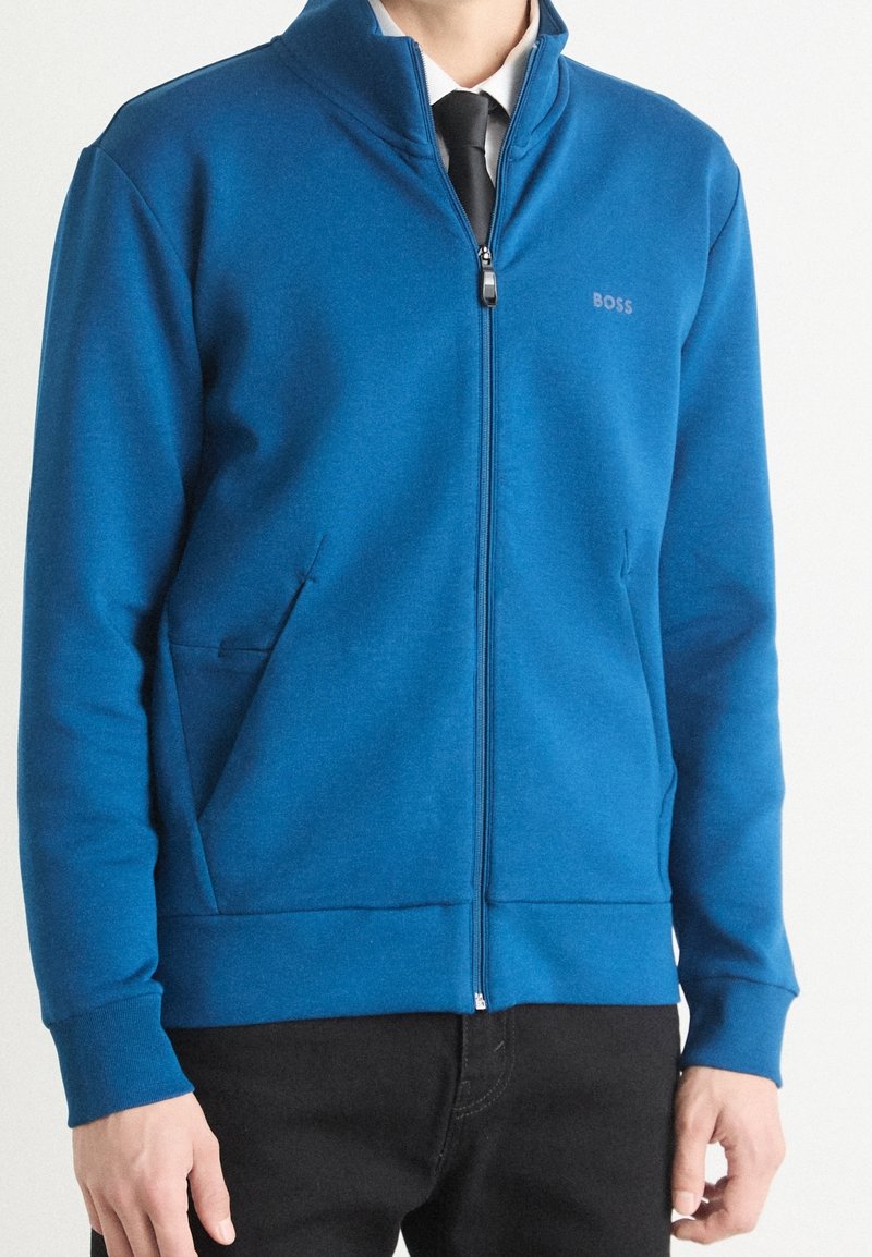 Blå zip-up jacka i mjukt tyg, med hög krage, två sidofickor och en diskret logotyp på bröstet.