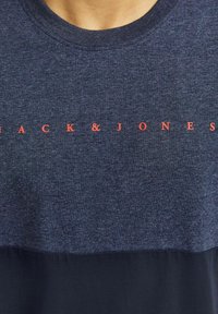 Tamsiai mėlynas marškinėliai su tekstūruotu paviršiumi, su raudonu "JACK & JONES" tekstu, centruotu krūtinės srityje. Apvalus iškirpimas, trumpomis rankovėmis.