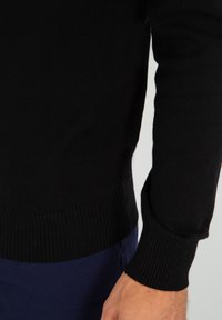 Maglione nero lavorato a maglia con polsini e orlo a coste, che mostra un primo piano del tessuto testurizzato e della finitura liscia, abbinato a pantaloni blu navy.
