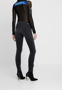 Kvinna i en svart genomskinlig långärmad topp med lappar och mörkgrå smala jeans, klädd i svarta högklackade ankelstövlar, ståendes.