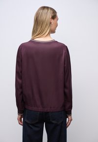 Chaqueta bomber de satén burdeos con puños y borde acanalados, que presenta un sencillo cuello redondo y una textura suave.