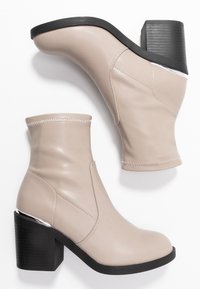 Botines beige con punteras redondeadas, tacones gruesos negros, detalles de costura y acentos plateados a lo largo de los bordes superiores del tacón.