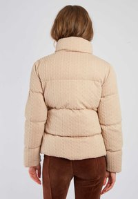 Kurtka puchowa w jasnobeżowym kolorze z teksturowanym wzorem dzianiny, wysokim kołnierzem i dopasowaną sylwetką. Posiada duże pikowane sekcje.