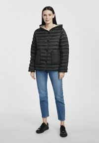 Czarna kurtka puchowa z poziomym przeszyciem, z zamykanymi kieszeniami i kapturem, w parze z niebieskimi dżinsami i czarnymi sneakersami.