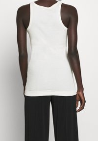 Valkoinen, hihaton tanktop, jossa on pehmeä rakenne ja pyöreä pääntie. Tässä on hienovarainen kontrastivärinen reunus käsivarsiaukkojen ja helman ympärillä.