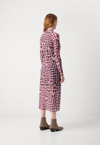 Lang, semi-transparant jurk met een roze en zwarte ruitpatroon, getailleerde lijfje, vloeiende rok en lange mouwen. Bruine enkelboots met lage hakken.