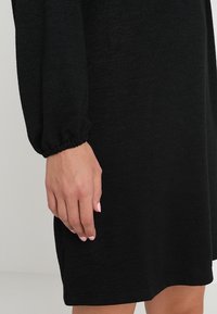 Vestido de punto negro con mangas largas. Los puños de las mangas son fruncidos y la tela parece suave con un acabado texturizado.