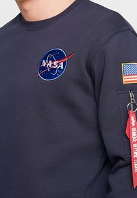 Felpa blu navy con il logo NASA e uno swoosh rosso, un toppe con la bandiera americana e una tasca laterale con un'etichetta rossa "RIMUOVERE PRIMA DEL Volo".