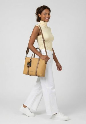 Donna sorridente che cammina, indossando un dolcevita beige senza maniche, pantaloni bianchi a gamba larga, scarpe da ginnastica bianche, porta una borsa a tracolla intrecciata color cuoio con un ciondolo di perline.