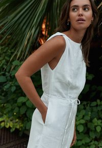 Robe en lin blanc avec un design sans manches, des poches latérales et une ceinture à la taille. Le tissu a une texture lisse, se détachant sur un fond de feuillage vert.