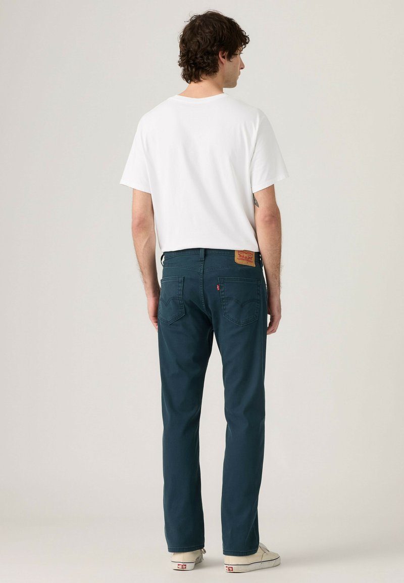 Mann trägt ein weißes T-Shirt mit kurzen Ärmeln, dunkelblaue Jeans und weiße Turnschuhe, steht mit dem Rücken zu einem schlichten hellen Hintergrund.