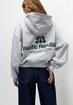 Personne aux cheveux longs porte un sweat à capuche gris avec le texte "Pacific Republic Youthful Design Collection Established 2014" et un jean bleu foncé, de dos.
