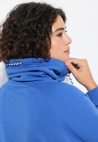 Blå sweatshirt med hög krage. Har en dragsko vid halsen och en texturerad yta. Synlig sömnad på ärmarna.