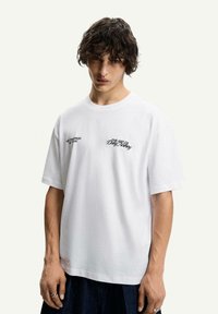 White cotton t-shirt with short sleeves, round neckline, featuring embroidered text in black on the chest. Simple, clean design.