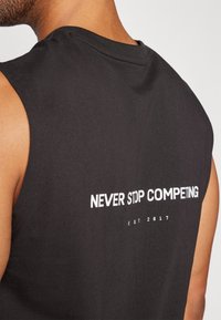Black sleeveless cotton shirt featuring white text on the back stating "NEVER STOP COMPETING" and "EST 2017." Smooth texture, casual fit.