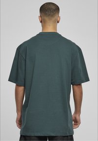 Uomo con capelli corti di spalle, indossa una t-shirt oversize a maniche corte di un verde scuro e pantaloni scuri su uno sfondo grigio uniforme.
