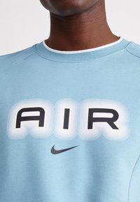 Ljusblå sweatshirt med ordet "AIR" i fet svart stil med vita vågiga konturer; inkluderar en liten svart Nike-logotyp.