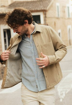 Mand iført beige jakke over lyseblå skjorte med knapper, justerer jakkefoer mens han står udendørs foran beige bygninger.