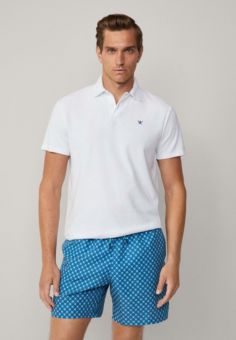Camisa polo blanca con mangas cortas y un pequeño logo azul marino, combinada con pantalones cortos de baño azules con un patrón de círculos. Textura suave, ajuste casual.