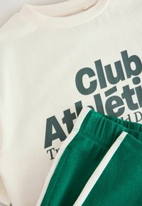 Camiseta de algodón crema con texto gráfico en verde oscuro, combinada con pantalones cortos de pana verdes con acentos blancos y textura acanalada.