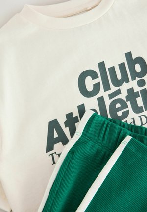 Kremowa bawełniana koszulka z grafitowym zielonym napisem, w połączeniu z zielonymi szortami z kordurowej tkaniny z białymi akcentami i prążkowaną teksturą.