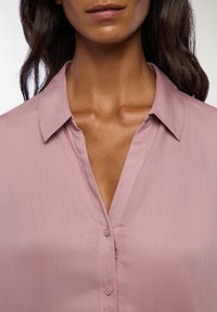 Camicia button-up rosa chiaro realizzata in tessuto liscio con colletto e frontale con bottoni. Presenta un scollo a V per un ulteriore dettaglio.