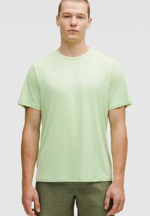 Junger Mann, der nach vorne steht, trägt ein hellgrünes kurzärmliges T-Shirt und strukturierte olivgrüne Hosen vor einem einfarbigen Hintergrund.