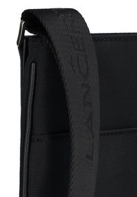 Czarny materiałowy pasek na ramię o teksturowanej powierzchni i wytłoczonym logotypie. Posiada metalowe elementy i wyraziste detale strukturalne.