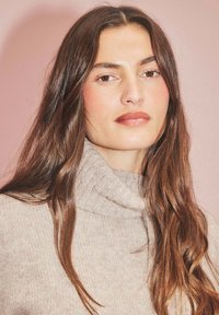 Hellbeige gestrickter Rollkragenpullover mit geripptem Muster. Das Modell hat lange braune Haare vor einem pinken Hintergrund. Neutrale Farbgestaltung.