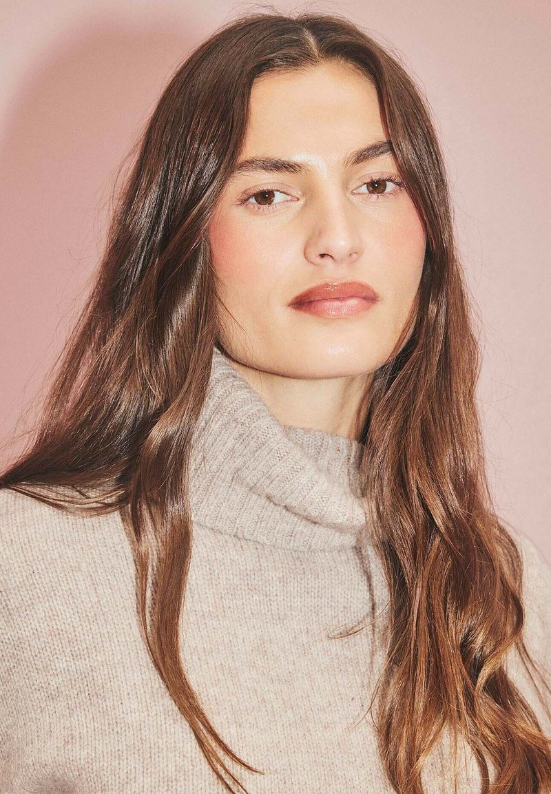 Hellbeige gestrickter Rollkragenpullover mit geripptem Muster. Das Modell hat lange braune Haare vor einem pinken Hintergrund. Neutrale Farbgestaltung.