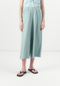 Culottes larges vert clair en tissu doux avec une taille élastique. Associés à des tongs noires sur pieds nus.